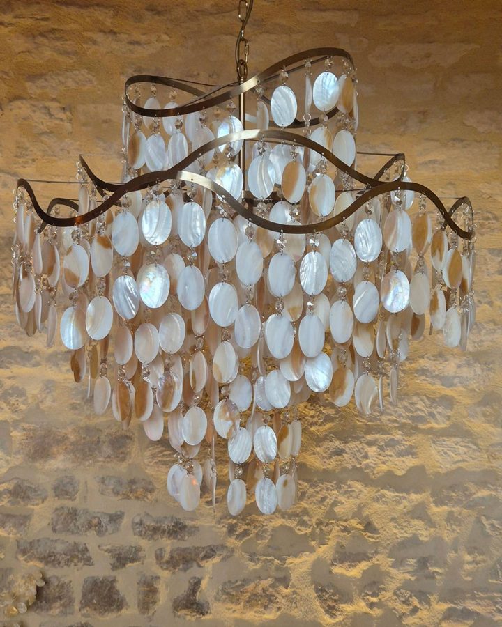 Lustre en nacre aux pampilles ovales irisées, suspendu devant un mur de pierre éclairé, au cabinet de réflexologie de Carine Laugier